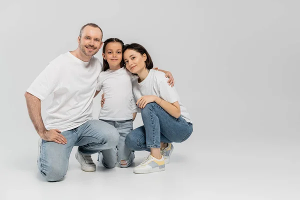 Happy father and mother with short hair hugging cheerful preteen daughter while sitting together in white t-shirts and blue denim jeans on grey background, Happy children's day, June 1 — Stock Photo