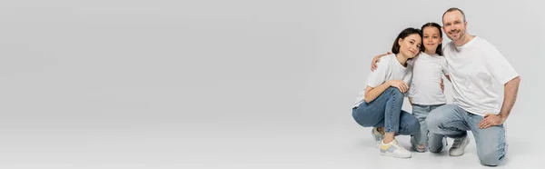 Allegro padre e madre con i capelli corti che abbracciano la figlia adolescente mentre si siedono insieme in t-shirt bianche e jeans in denim blu su sfondo grigio, Giornata per la protezione dei bambini, banner — Foto stock