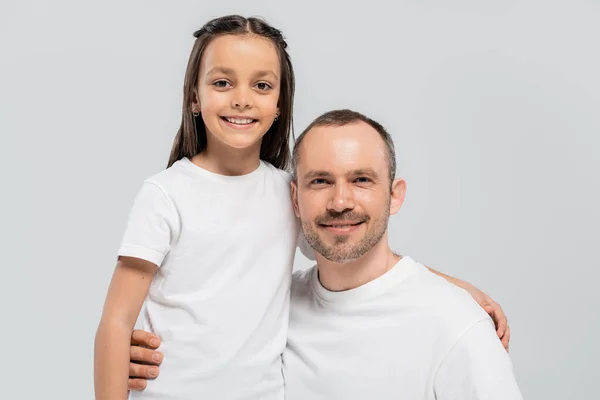 Unshaved and cheerful father with bristle hugging happy preteen daughter with long brunette hair while posing in white t-shirts and looking at camera on grey background, Child protection day — Stock Photo
