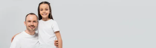 Unshaved and cheerful father with bristle hugging happy preteen daughter with long brunette hair while posing in white t-shirts and looking at camera on grey background, Child`s protection day, banner — Stock Photo