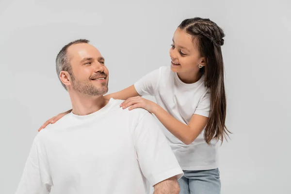 Счастливая девочка-подросток с длинными волосами брюнетки опираясь на небритый и веселый отец с щетиной во время позирования в белых футболках и глядя друг на друга на сером фоне, День защиты детей — стоковое фото