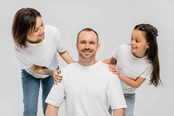 Женщина с короткими волосами и татуировкой на руке и брюнетка-подросток в джинсах в голубой джинсе, смотрящая на мужчину в белой футболке на сером фоне, счастливая семья — стоковое фото