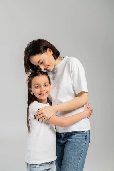 Glückliche Mutter mit kurzen Haaren und Tätowierung an der Hand, die ihre Tochter umarmt, während sie in weißen T-Shirts und blauen Jeans vor grauem Hintergrund zusammensteht, Internationaler Kinderschutztag — Stockfoto
