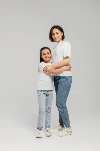 Full length of mother with short hair and tattoo on hand hugging preteen daughter while standing together in white t-shirts and blue denim jeans on grey background, International child protection day — Stock Photo