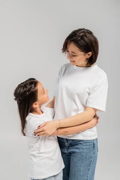Happy mother with short hair and tattoo on hand hugging brunette daughter while standing together in white t-shirts and blue denim jeans on grey background, International child protection day — Stock Photo