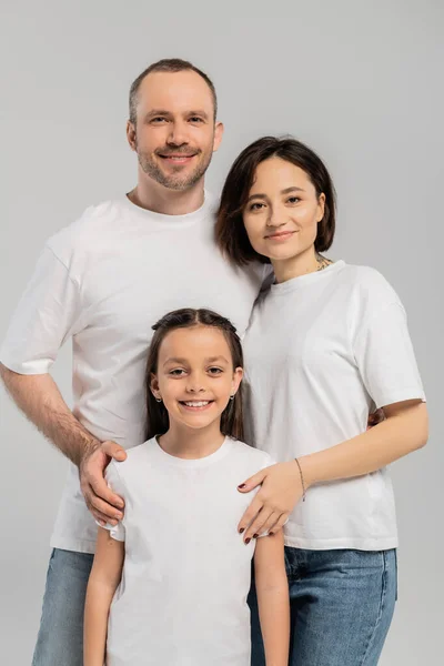 Retrato de familia feliz en camisetas blancas mirando a la cámara sobre fondo gris, Día Internacional de la Protección del Niño, padre y madre con tatuaje abrazando a la hija morena preadolescente - foto de stock