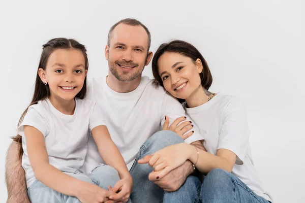 Retrato de família feliz em camisetas brancas e jeans jeans jeans azul olhando para a câmera e sentados juntos no fundo cinza, Dia Internacional de Proteção à Criança, pais e filha — Fotografia de Stock