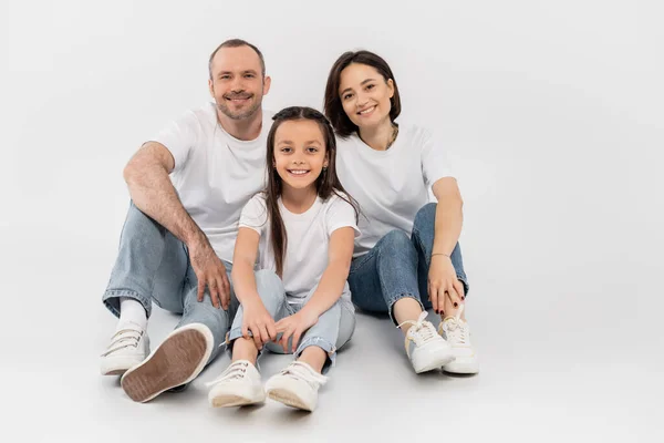 Стильная семья в белых футболках и джинсах в синих джинсах, смотрящая на камеру и сидя вместе на сером фоне, Международный день защиты детей, счастливые родители и дочь — стоковое фото