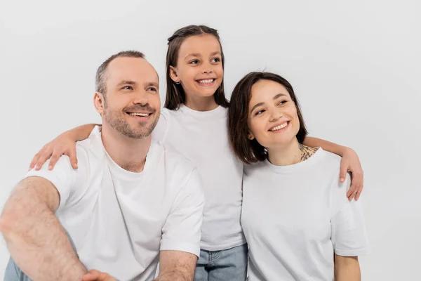 Familia con estilo en camisetas blancas mirando hacia otro lado mientras posan juntos sobre un fondo gris, Día Internacional de la Protección del Niño, hija preadolescente abrazando a padres felices - foto de stock