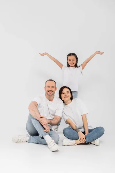 Preteen daughter standing with outstretched hands near happy parents in white t-shirts and blue denim jeans looking at camera and posing together on grey background, child protection day — Stock Photo