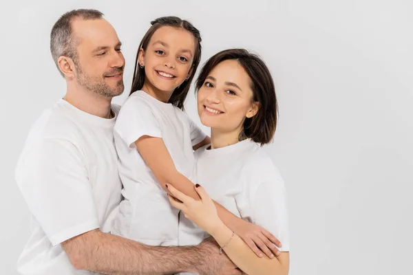 Портрет счастливой семьи в белых футболках, смотрящей в камеру и обнимающей друг друга на сером фоне, Международный день защиты детей, родители и дочь — стоковое фото