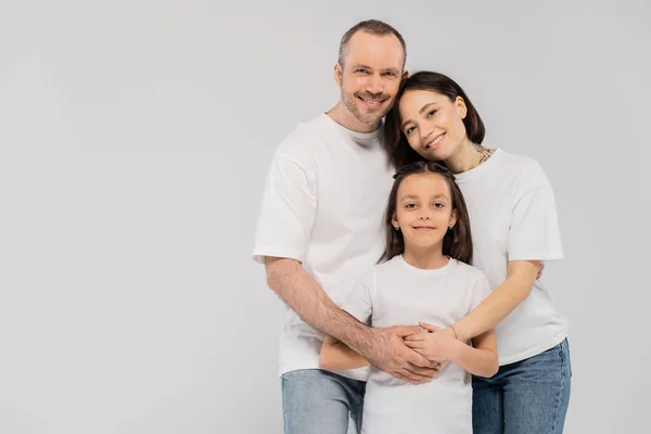 Счастливые родители обнимают веселую дочь, стоя вместе в джинсах в синих джинсах и белых футболках и глядя на камеру на сером фоне, счастливого детского дня, 1 июня — стоковое фото