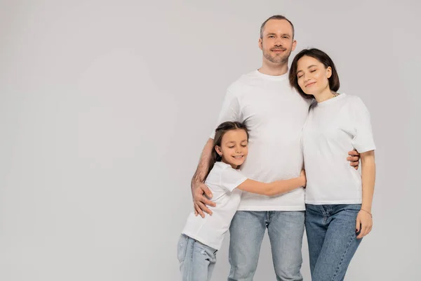 Happy preteen girl hugging father near mother with tattoo while standing together in blue denim jeans and white t-shirts and looking at camera on grey background, Happy children's day — Stock Photo