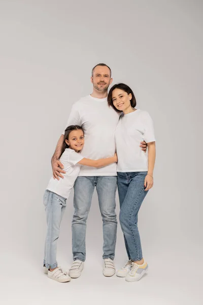Joyous preteen girl hugging father near mother with tattoo while standing together in blue denim jeans and white t-shirts and looking at camera on grey background, Happy children's day, — Stock Photo