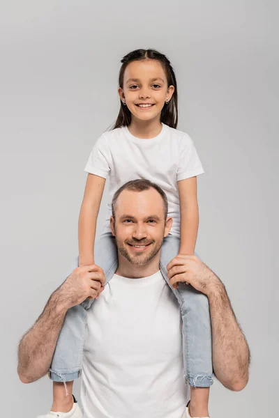 Happy preteen daughter with brunette hair sitting on shoulders of unshaved and cheerful father with bristle and looking at camera on grey background, Child protection day — Stock Photo
