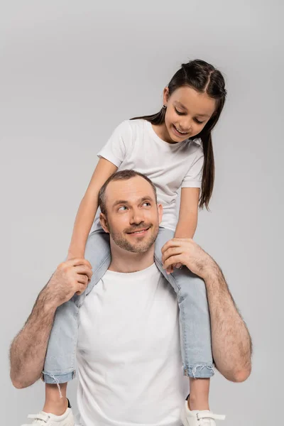 Feliz hija preadolescente con el pelo largo morena sentado en los hombros de padre sin afeitar y alegre con cerdas sobre fondo gris, Día de la protección del niño, padre y la hija - foto de stock