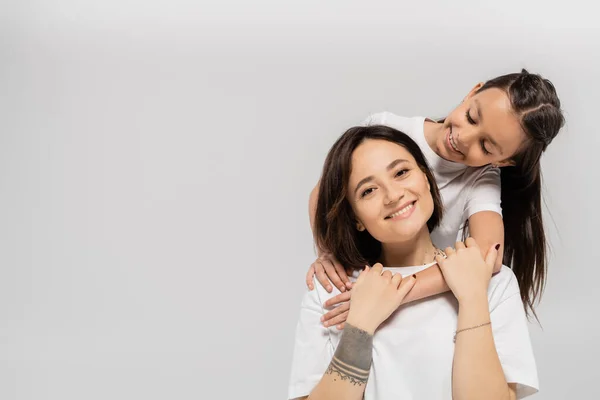 Happy preteen daughter with brunette hair embracing cheerful mother with tattoo on hand and looking at camera on grey background, Child protection day, mother and daughter — Stock Photo