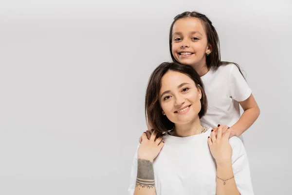 Happy preteen daughter with long brunette hair holding hands of cheerful mother with tattoo and looking at camera on grey background, Child protection day, mother and daughter — Stock Photo