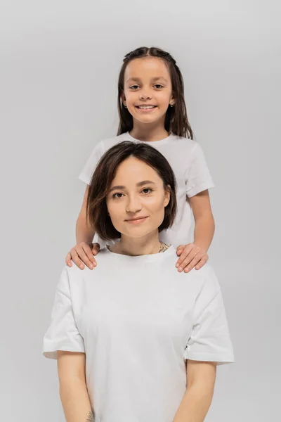 Happy preteen daughter with long brunette hair holding hands of cheerful mother with tattoo and looking at camera on grey background, mother and daughter, Happy children's day — Stock Photo