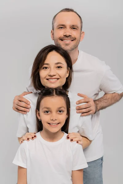 Famille heureuse en t-shirts blancs regardant la caméra et posant ensemble sur fond gris, Journée internationale de la protection de l'enfance, parents positifs et fille — Photo de stock