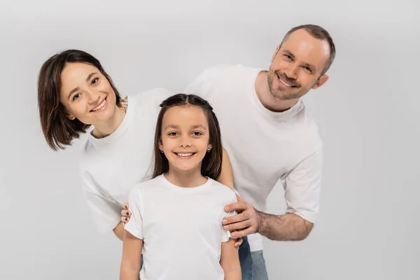 Familia feliz en camisetas blancas mirando a la cámara y posando juntos sobre fondo gris, Día Internacional de la Protección del Niño, padre positivo madre e hija - foto de stock