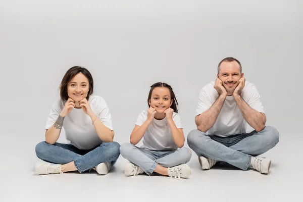 Joyous father and tattooed mother with short hair and cheerful preteen daughter sitting with crossed legs in white t-shirts and blue denim jeans on grey background, Happy children's day — Stock Photo