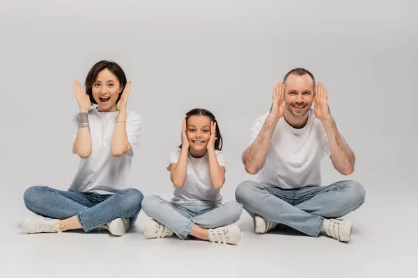 Amazed father and tattooed mother with short hair and excited preteen daughter sitting with crossed legs in white t-shirts and blue denim jeans on grey background, Happy children's day — Stock Photo