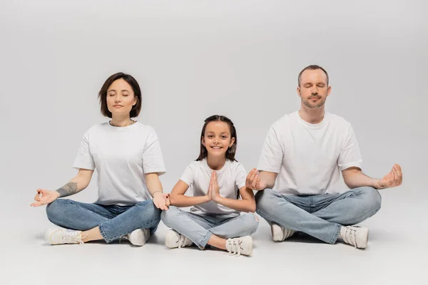 Father and tattooed mother with short hair meditating with cheerful preteen daughter while sitting with crossed legs in white t-shirts and blue denim jeans on grey background — Stock Photo