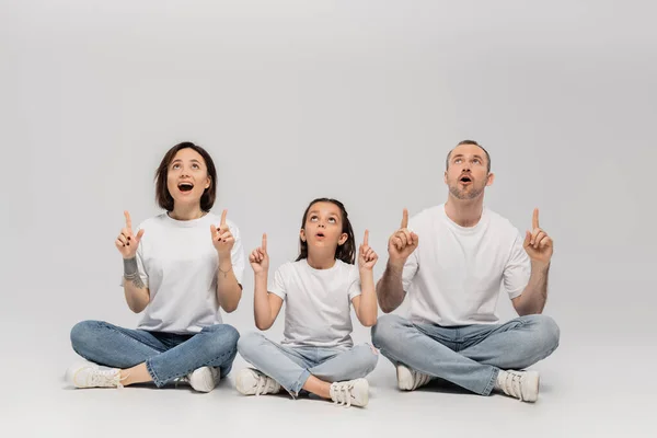 Amazed family sitting with crossed legs in white t-shirts and blue denim jeans while pointing with fingers and looking up on grey background, father, tattooed mother and preteen daughter — Stock Photo