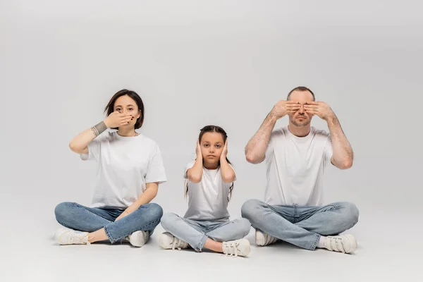 Família sentada com pernas cruzadas em camisetas brancas e jeans jeans jeans azul enquanto demonstra a sabedoria dos três macacos sábios; não veja mal, não diga mal e não ouça mal no fundo cinza — Fotografia de Stock