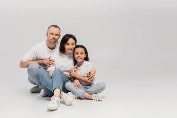 Pai alegre e mãe com cabelo curto abraçando alegre filha pré-adolescente enquanto sentado com pernas cruzadas em camisetas brancas e jeans jeans jeans azul no fundo cinza, dia feliz das crianças — Fotografia de Stock