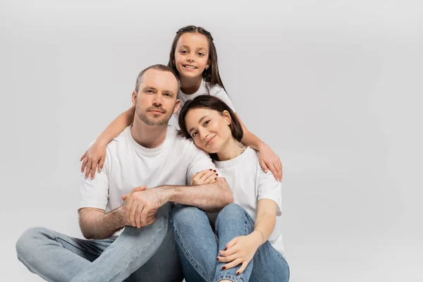 Joyous preteen girl hugging cheerful parents in white t-shirts and blue denim jeans while bonding together and looking at camera on grey background, Happy children's day — Stock Photo