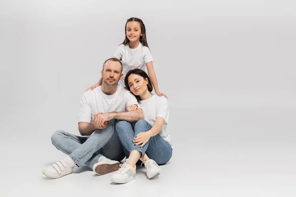 Fröhliches Mädchen, das Mutter und Vater in weißen T-Shirts und blauen Jeans umarmt, während es sich zusammenklebt und in die Kamera auf grauem Hintergrund blickt, Happy Children 's Day — Stockfoto