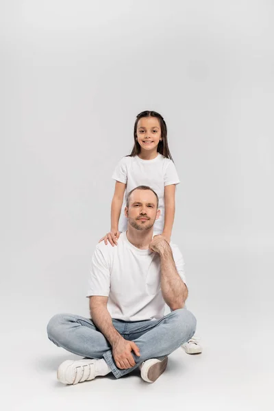 Happy preteen girl with brunette hair hugging cheerful father with bristle while posing in white t-shirts and blue denim jeans and looking at camera on grey background, Happy Father`s Day — Stock Photo