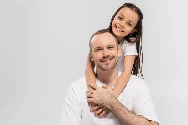 Portrait of happy daughter with long brunette hair hugging cheerful father with bristle while posing in white t-shirts and looking at camera on grey background, Happy Father`s Day — Stock Photo