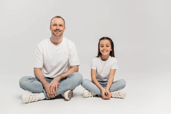 Happy preteen girl with long brunette hair and cheerful dad with bristle sitting with crossed legs while posing in white t-shirts and blue denim jeans on grey background, Happy Father`s Day — Stock Photo