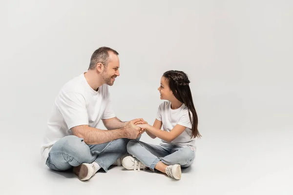 Happy preteen girl with long brunette hair holding hands with cheerful dad while sitting with crossed legs in white t-shirts and blue denim jeans on grey background, Happy Father`s Day — Stock Photo