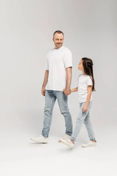 Ein fröhlicher Vater mit Borsten und eine frühpubertäre Tochter in weißen T-Shirts und blauen Jeans, die sich an den Händen halten und gemeinsam auf grauem Grund spazieren gehen. — Stockfoto