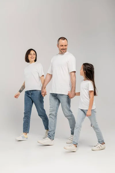Full length of cheerful parents and preteen daughter in white t-shirts and blue denim jeans holding hands and walking together on grey background, International child protection day in June — Stock Photo