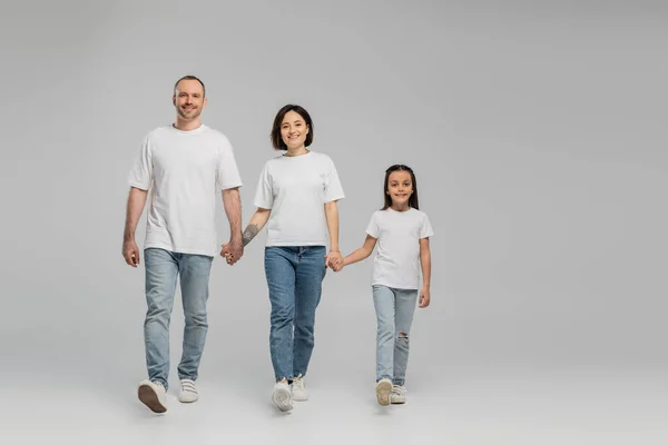 Full length of happy parents and daughter in white t-shirts and blue denim jeans holding hands and walking together on grey background, International child protection day in June — Stock Photo