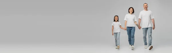 Pleine longueur de parents heureux et fille en t-shirts blancs et jeans en denim bleu tenant la main et marchant ensemble sur fond gris, Journée internationale de la protection de l'enfance en Juin, bannière — Photo de stock