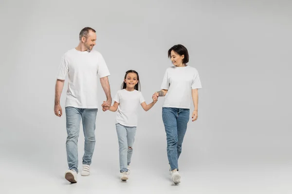 Full length of happy parents and kid in white t-shirts and blue denim jeans holding hands and walking together on grey background, International child protection day in June — Stock Photo