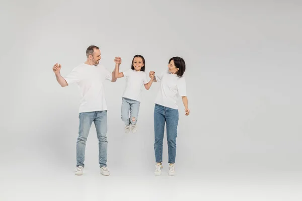 Полная длина счастливых родителей в белых футболках и джинсах в синих джинсах, держащихся за руки и поднимающих дочь-подростка, стоя вместе на сером фоне, Международный день защиты детей в июне — стоковое фото