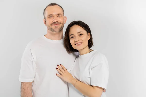 Cheerful and tattooed woman with short brunette hair hugging joyous husband with bristle while standing together in white t-shirts and looking at camera isolated on grey background, happy couple — Stock Photo
