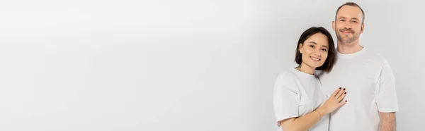 Татуйована жінка з коротким волоссям брюнетки обіймає щасливого чоловіка з щетиною, стоячи разом у білих футболках і дивлячись на камеру ізольовано на сірому фоні, щаслива пара, банер — стокове фото