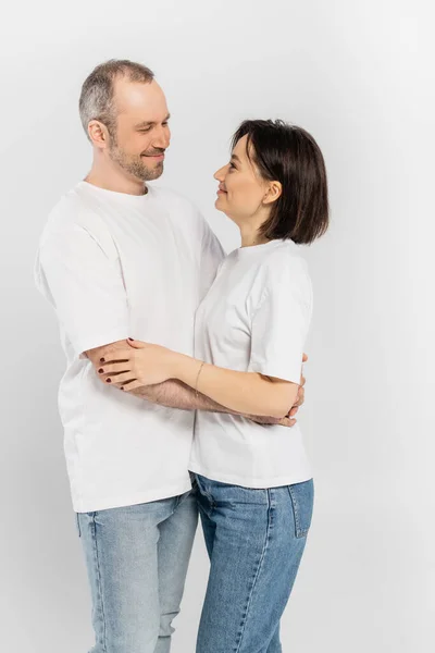 Mujer alegre y tatuada con el pelo corto morena abrazando alegre marido con cerdas mientras están de pie juntos en camisetas blancas y mirándose aislados sobre fondo gris, feliz pareja - foto de stock