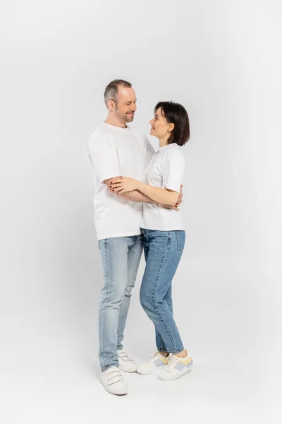 Full length of smiling woman with short brunette hair hugging husband with bristle while standing together in white t-shirts and denim jeans looking at each other on grey background, happy couple — Stock Photo
