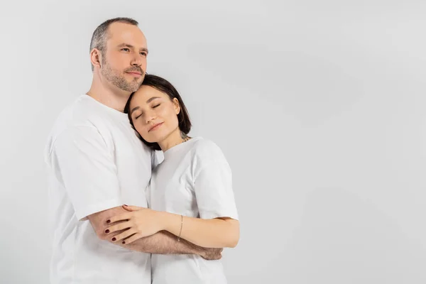 Mujer tatuada con el pelo corto morena y los ojos cerrados apoyados en el pecho del marido con cerdas mientras están de pie juntos en camisetas blancas aisladas sobre fondo gris, feliz pareja - foto de stock