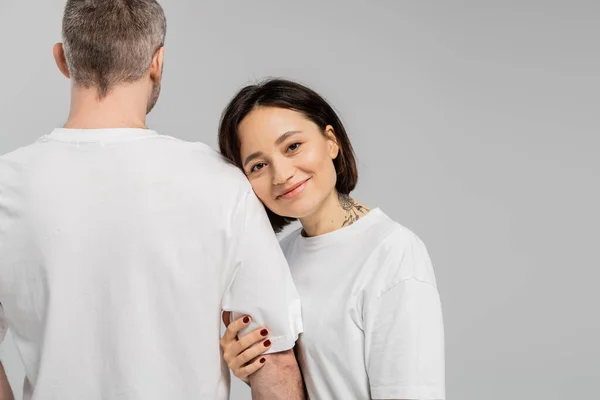 Cheerful and tattooed woman with short brunette hair leaning on shoulder of husband while standing together in white t-shirts and looking at camera isolated on grey background, happy couple — Stock Photo