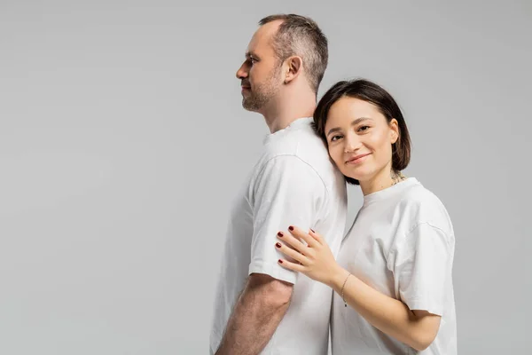 Tattooed woman with short brunette hair leaning on back of husband while standing together in white t-shirts and looking at camera isolated on grey background, happy couple — Stock Photo